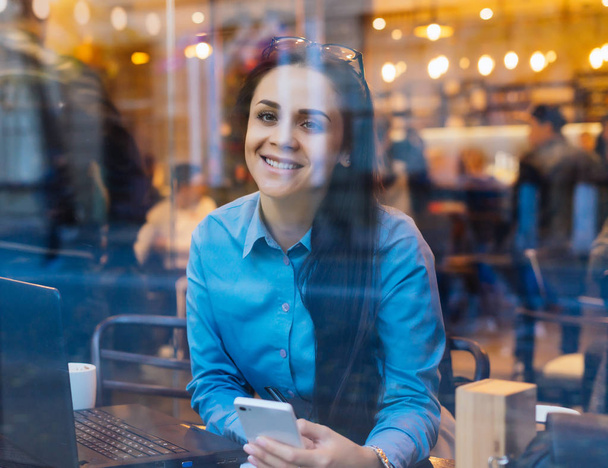 heureux jeune fille indépendante réussie assis dans le café par la fenêtre, travaillant sur ordinateur portable et smartphone
 - Photo, image