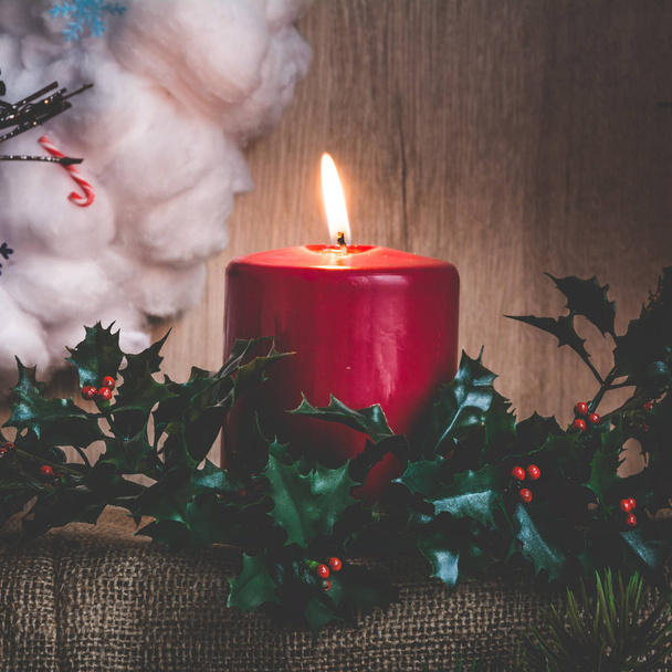Горящая свеча и ветви падуба на фоне рождественского венка из ваты
 - Фото, изображение