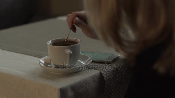 Young woman mixing sugar in coffee cup on table on background mobile phone - Záběry, video