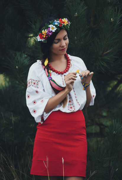 Beautiful woman in colorful ukrainian traditional dress holding herself and enjoying summer in green forest - Photo, Image