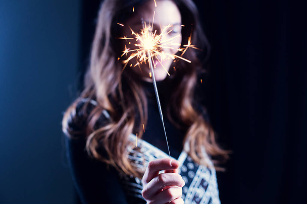 Happy girl smiling and holding a sparkling firework in her hand. Girl on the background intentionally blured. The concept of celebrating the New Year and Christmas. - Foto, Imagen