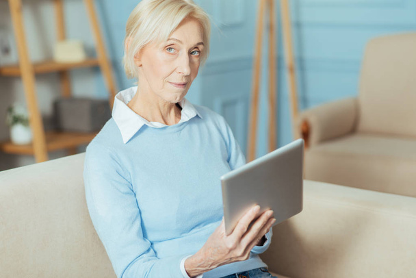 Curious aged woman sitting on a sofa and holding a modern tablet - Fotó, kép