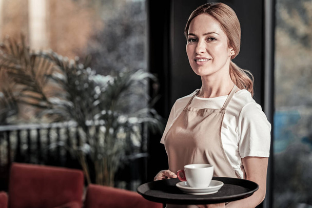 Calm friendly waitress standing with a cup of coffee on her tray - Zdjęcie, obraz