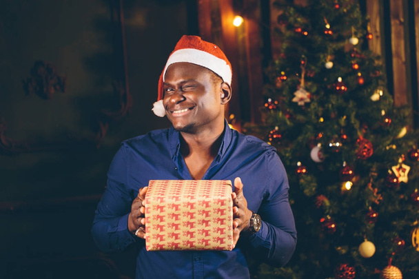 Afro amerykański facet z uroczym uśmiechem trzymając w rękach prezent na Boże Narodzenie - Zdjęcie, obraz
