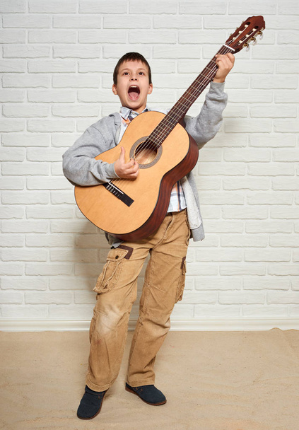 chłopiec odtwarzanie muzyki na gitarze, stały w pełnej wysokości, biały cegła tle muru - Zdjęcie, obraz