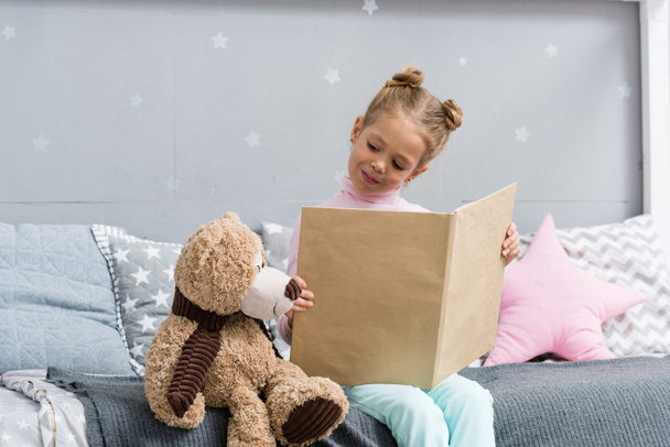adorable little kid reading book in bed with teddy bear - Fotó, kép