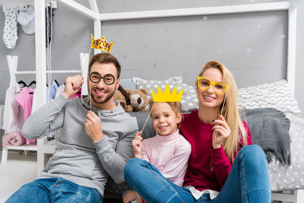 happy young family with masquerade crowns and eyeglasses sitting in kid bedroom - Fotó, kép