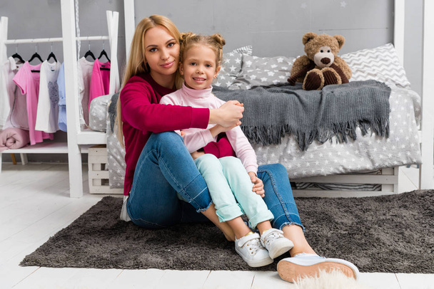 young mother and daughter embracing while sitting on floor of kid bedroom - Photo, image