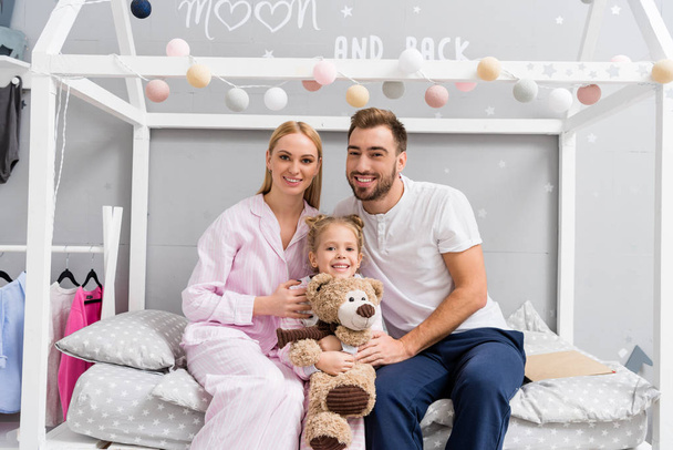 улыбающаяся молодая семья, сидящая на кровати в детской спальне и смотрящая в камеру
 - Фото, изображение