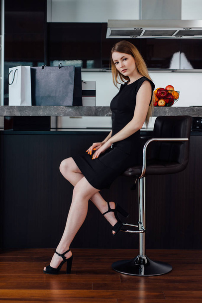 Полноразмерный портрет элегантной женщины со светлыми волосами в черном платье и на высоких каблуках, сидящей на барном стуле на кухне
 - Фото, изображение