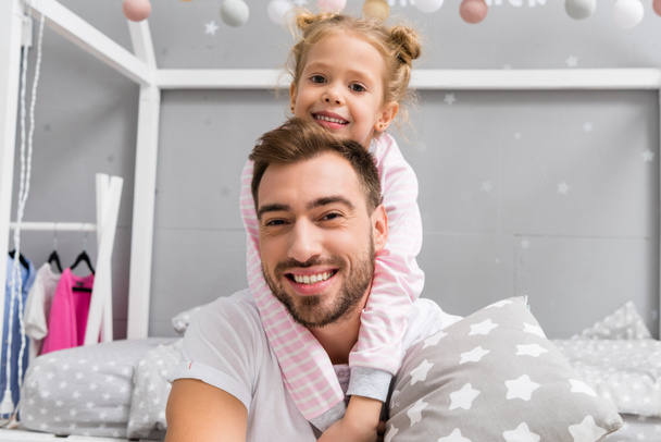 happy little daughter embracing father from behind in kid bedroom - Фото, изображение