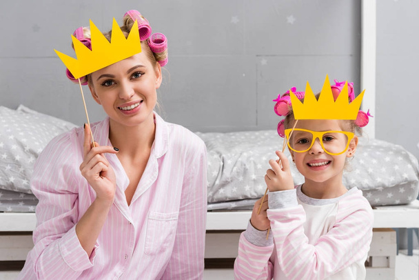 smiling mother and daughter in pajamas with toy masquerade crowns - Фото, изображение