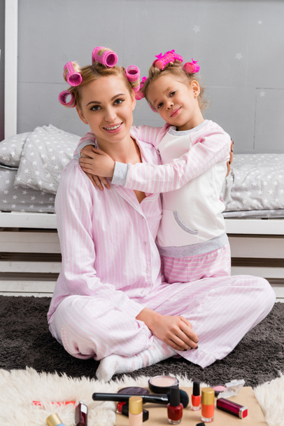 happy mother and daughter with hair rollers and in pajamas sitting on floor - Fotoğraf, Görsel