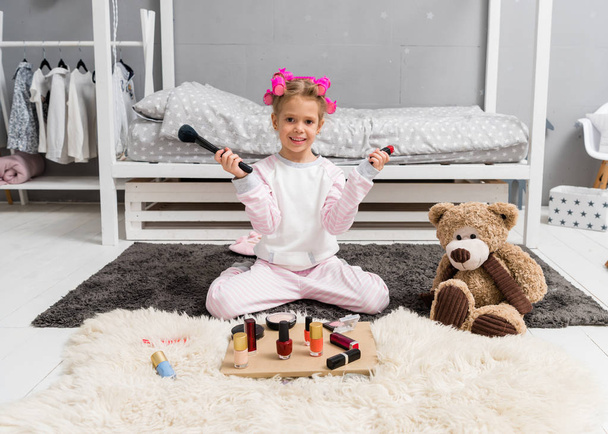 adorable little kid with hair rollers on head and makeup tools sitting on floor - Φωτογραφία, εικόνα