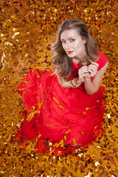 Beautiful young woman with stylish haircut wearing red dress, sitting on golden studio background surrounded by golden confetti, holiday celebration concept - Zdjęcie, obraz