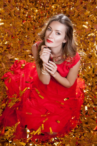Krásná mladá žena s stylový účes na sobě červené šaty, sedí na golden studio pozadí obklopen zlaté konfety, dovolená oslava concept - Fotografie, Obrázek