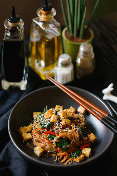 glass noodles with seasonal vegetables on black plate with chopsticks and olive oil, balsamic sauce, salt, pepper, vase on background - Photo, Image