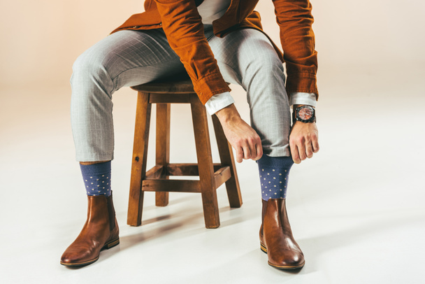 cropped shot of stylish man tying socks while sitting on wooden chair, on beige - Photo, image