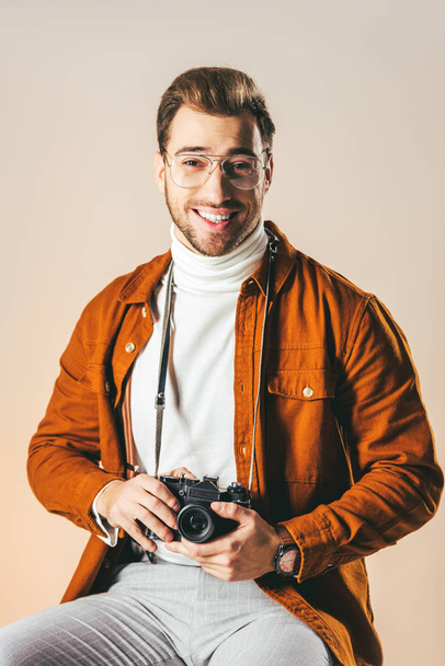 Porträt eines fröhlichen Mannes in stylischer Kleidung mit Fotokamera, der isoliert auf beige blickt - Foto, Bild