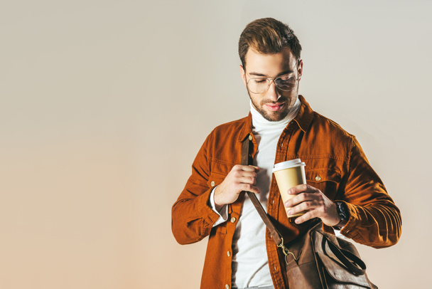 portrait of fashionable man with bag looking at disposable cup of coffee in hand isolated on beige - Photo, Image