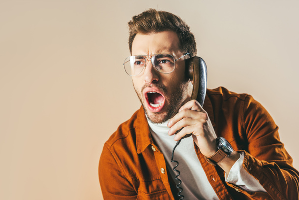 portrait of stylish man screaming while talking on telephone isolated on beige - 写真・画像