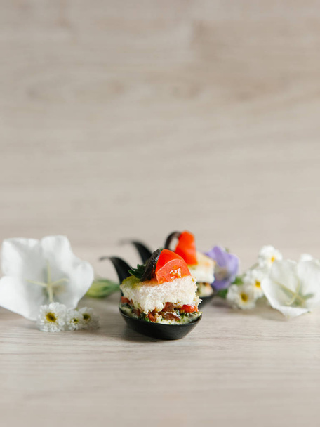 Weinig canapeetjes in keramische lepels versierd met tomaat slice en olijf ringen, met bloemen op houten achtergrond - Foto, afbeelding