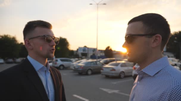 Dwóch młodych biznesmenów stojących na zewnątrz przy parkingu i rozmawiających. Profil biznesmenów w okularach przeciwsłonecznych przemawiających na zewnątrz miasta. Komunikacja szczęśliwych kolegów. Zamknij Powolny ruch - Materiał filmowy, wideo