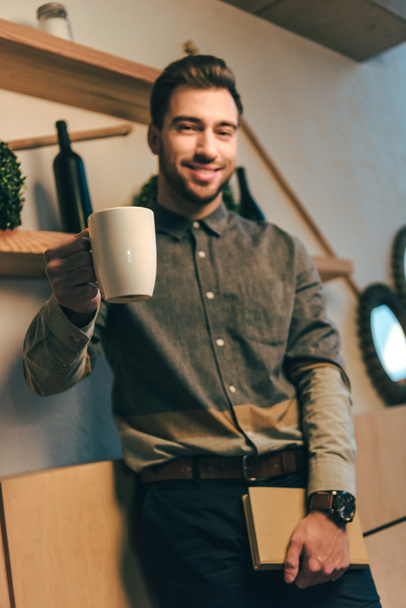 selectieve aandacht van glimlachende man met kopje koffie en een boek in handen in café - Foto, afbeelding