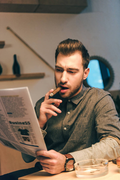portrait of confident businessman smoking cigar while reading newspaper in cafe - Photo, Image
