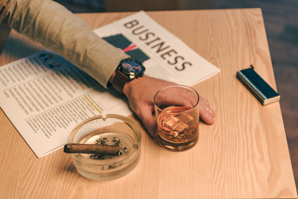 περικοπεί shot του επιχειρηματία που κάθεται στο τραπέζι με ποτήρι κονιάκ, εφημερίδα και πούρο στο τασάκι στο καφέ - Φωτογραφία, εικόνα