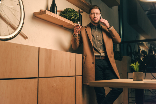 portrait of stylish man in jacket with glass of whiskey talking on smartphone in cafe - Zdjęcie, obraz