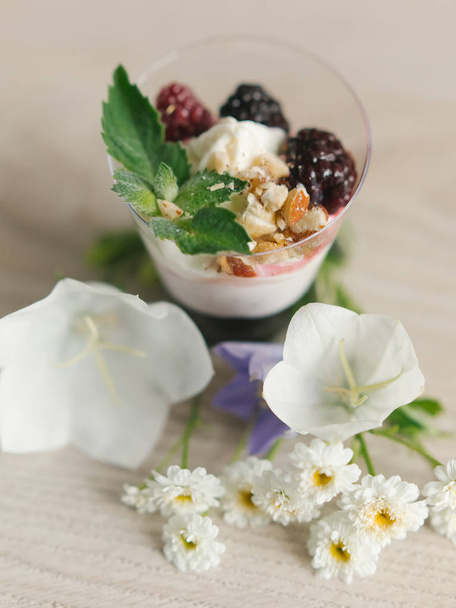 Yogur griego con mermelada de mora y nueces, menta y moras en la parte superior, con flores alrededor en la superficie de madera
   - Foto, Imagen