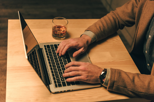Oříznout záběr člověka psaní na notebooku u stolu se sklenkou whisky - Fotografie, Obrázek
