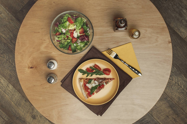 top view of piece of pizza on plate, salad in bowl and spices on wooden surface - Фото, изображение