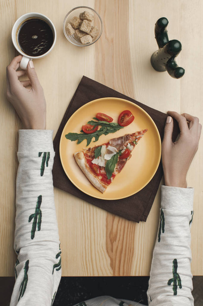 partial view of woman sitting at table with pizza piece on plate and cup of coffee - Foto, Imagem