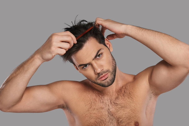 Good looking young man combing his hair while standing against grey background - Foto, immagini