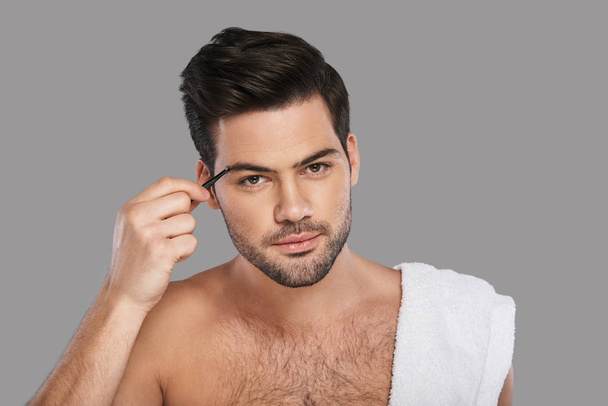 Handsome young man applying eyebrow tweezers against grey background - Photo, image