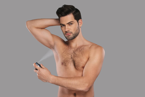 shirtless handsome young man applying deodorant while standing against grey background - 写真・画像