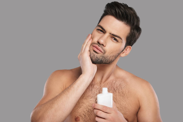 Handsome young man applying aftershave lotion and standing against grey background - Fotoğraf, Görsel