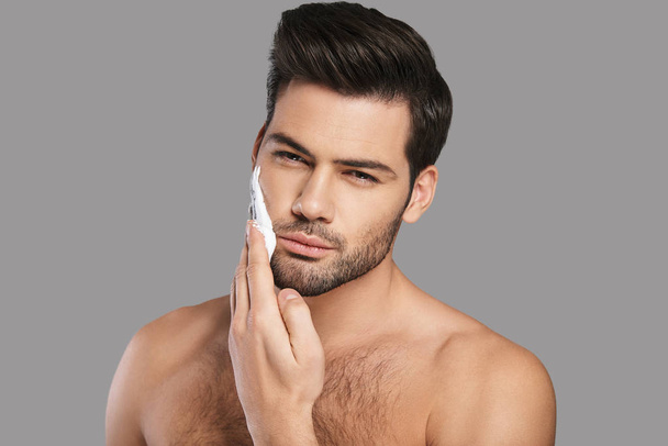 Hombre joven guapo aplicando crema de afeitar y mirando a la cámara sobre fondo gris
 - Foto, imagen