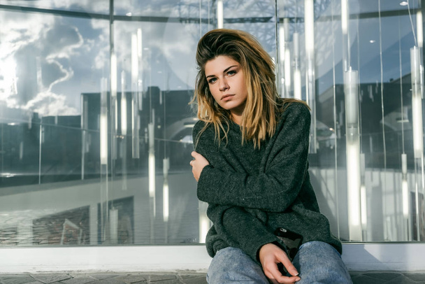 sad stylish girl with thick hair sitting near a glass wall - Photo, Image