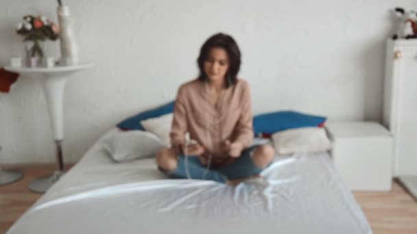 charming brunette woman sits on the bed and dancing to music in headphones a blurred background - Séquence, vidéo