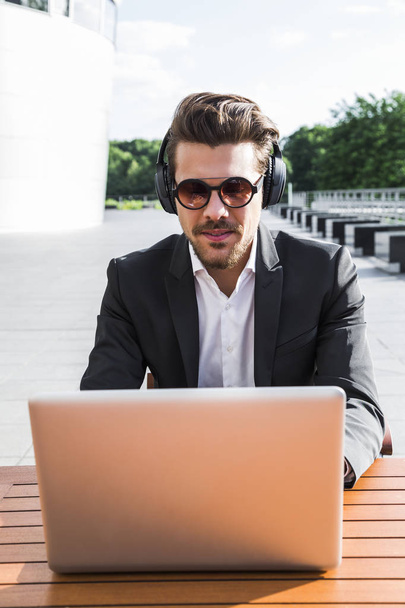 businessman in sunglasses, black suit with shirt sitting outside at wooden orange table at cafe and typing and working on computer and listening to music in headphones - Foto, Bild