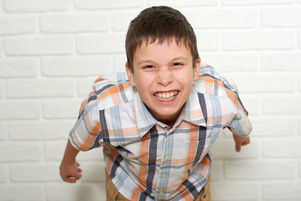 portrait of a emotional boy standing near white brick wall, dressed in a plaid shirt - Photo, Image