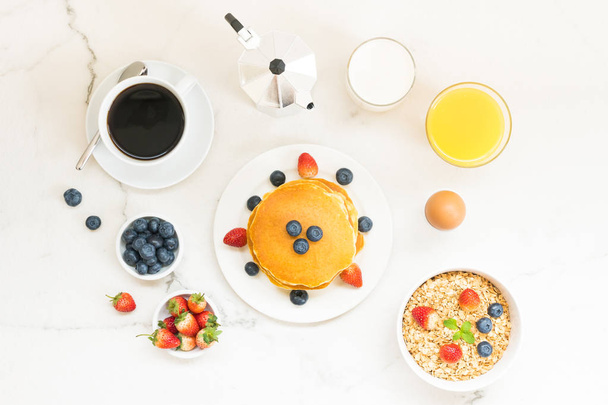 gesundes Frühstücksset mit Pfannkuchen und Müsli mit Blaubeere und Erdbeere und schwarzem Kaffee, Milch und Orangensaft auf weißem Steintischhintergrund - Foto, Bild