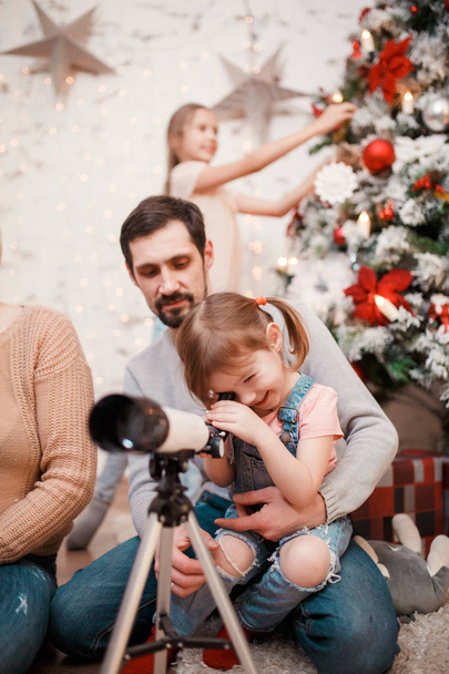 Image of parents and daughters with telescope on background of decorated New Years fir - Photo, Image