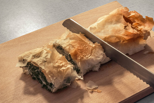 Rolo de torta de espinafre de queijo sérvio tradicional recém-assado cortado com faca de pão serrilhada na placa de corte de madeira
 - Foto, Imagem