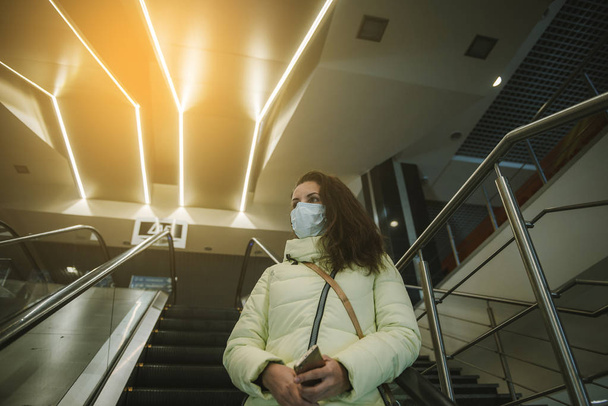 Persoon draagt beschermend masker tegen overdraagbare infectieziekten en als bescherming tegen verontreiniging en de griep. Vrouw in een winkelcentrum op een roltrap - Foto, afbeelding