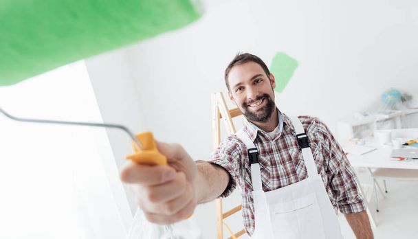 Smiling professional painter working and painting, paint roller on the foreground, home renovation and decoration concept - Fotoğraf, Görsel