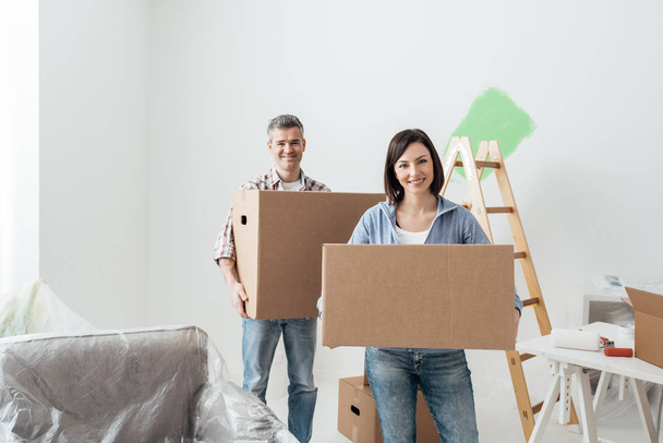 Happy couple moving into a new house and doing home renovations, they are carrying cardboard boxes - Foto, Imagem